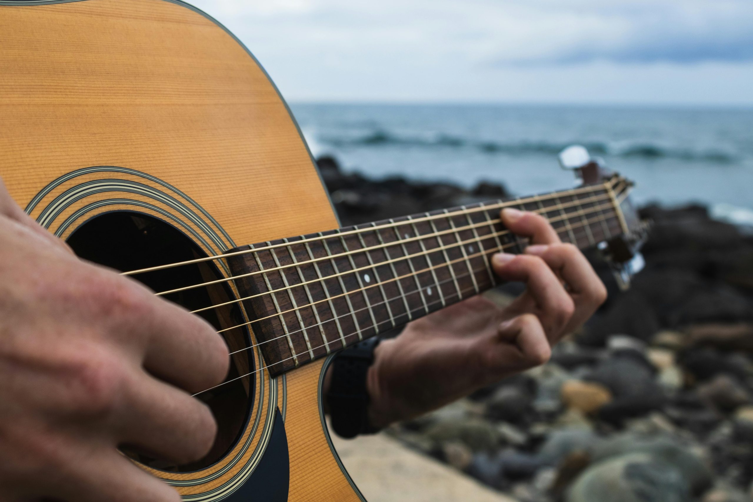 Close-up of hands playing an acoustic guitar on a rocky beach with a view of the ocean.